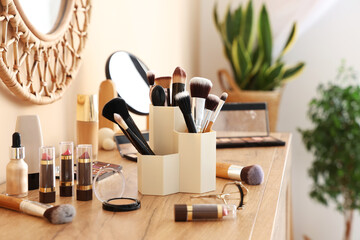 Holder with makeup brushes and different decorative cosmetics on chest of drawers at home