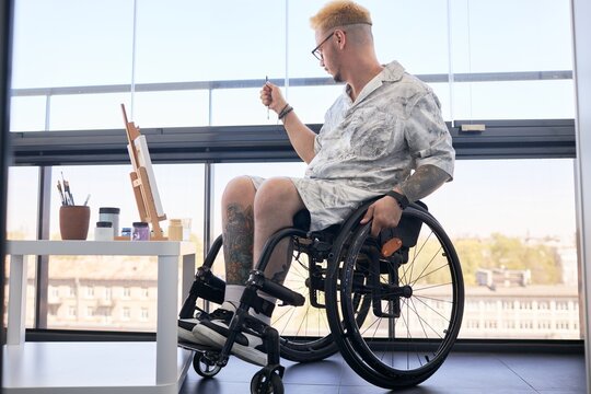 Man with tattoos, sitting in wheelchair, painting on easel in bright room with large windows and city view, creating art piece in casual attire with focus on creativity and concentration