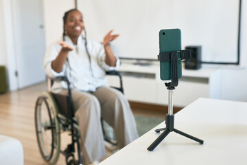 Fototapeta premium Smiling woman in wheelchair communicating using smartphone on tripod in living room with modern decor, expressing joy and engagement