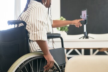 Woman in wheelchair engaged in virtual meeting using smartphone on tripod while sitting in cozy room with bright light and decor on background