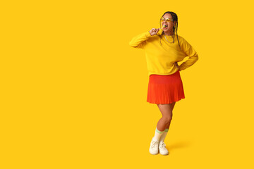 Beautiful young African-American woman with dreadlocks holding lollipop on yellow background