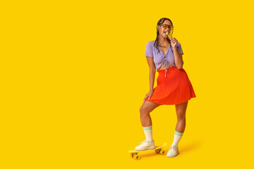 Beautiful young African-American woman with dreadlocks holding lollipop and skateboard on yellow background