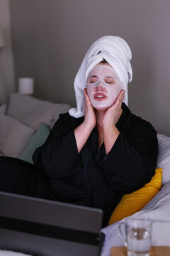Plus size woman in pajamas using facial mask in front of laptop