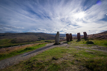 The image is of a group of rocks in a field,  part of a landscape in Scotland known as The Watchers on the Snow Roads. The stones are set against a backdrop of grass, sky, and mountains.