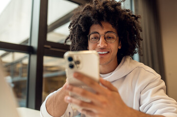 Male student checking mobile phone sitting at desk in library campus using laptop studying online