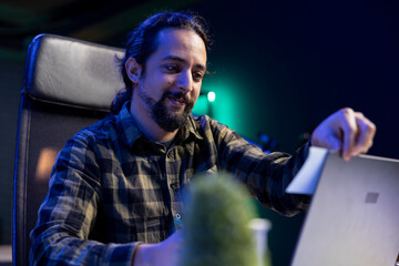 Male university student studying with a laptop and utilizing online resources focused on academic research and learning, surrounded by notes and textbooks. Man placing a sticky note on device screen.