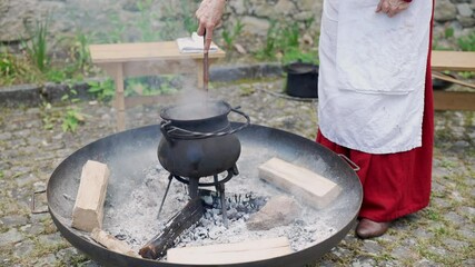 Person stirring a boiling cauldron over an open fire during a medieval reenactment, showcasing traditional outdoor cooking methods and the authentic feel of the historical event
