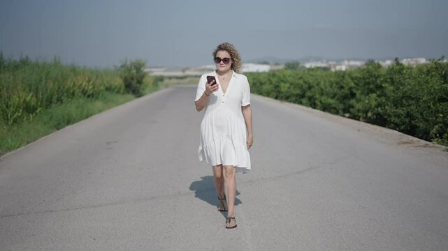 young curly-haired blonde Caucasian woman in a short white dress walking facing the camera on a lonely road in the outskirts answers a call from her smartphone
