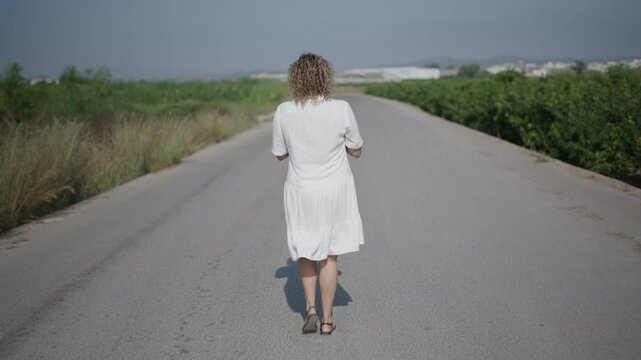 young girl curly haired blonde Caucasian woman in a short white dress walking with her back to the camera gathers her hair with a hair clip