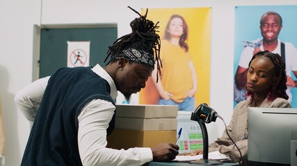 African american guy picking up order from clothing store, signing confirmation papers at cash register. Woman employee preparing boxes with requested fashion items and accessories. Camera A.