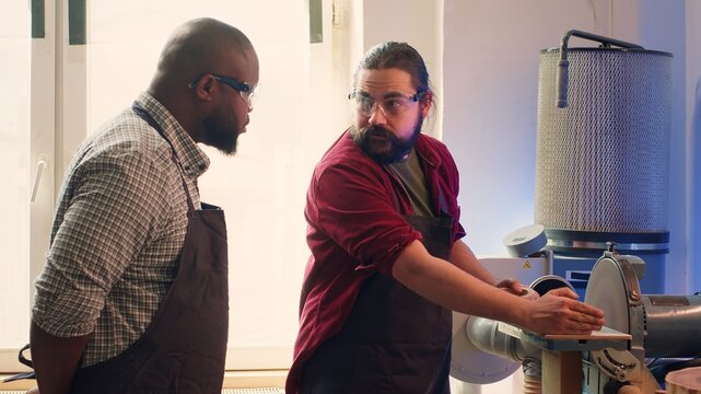 Manufacturer teaching new employee how to shape wooden bowl on disc sander in assembling shop. Apprentice learning from woodworking specialist how to use grinding machine rotating disc, camera A