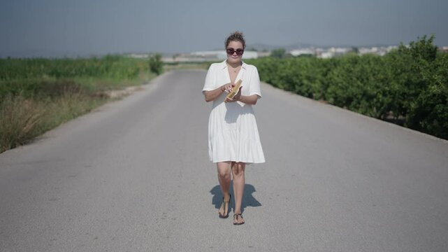 Young curly haired blonde Caucasian woman in a short white dress walking facing the camera on a lonely suburban road reading a book