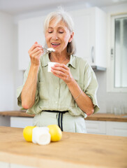 Positive elderly woman eating tasty yogurt at kitchen interior, healthy snack