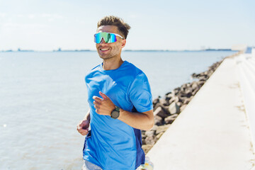 A man in a blue shirt runs along a seaside path, smiling under the bright sun with reflective sunglasses.