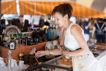 Woman in flea market store look at crystal glass, antique gifts. Female buyer holds item in hands...