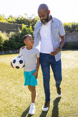 Naklejka premium Grandfather and grandson walking on grass field, holding soccer ball, enjoying time together