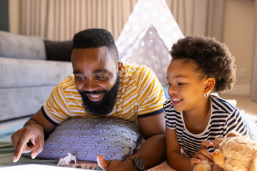 Father and daughter lying on floor, using tablet and smiling at home, in teepee