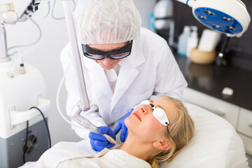 Fototapeta premium Focused skilled female cosmetologist performing hardware laser treatment to resurfacing and tightening facial skin to young woman at cosmetology clinic
