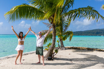 Happy married couple raise their arms in victory and celebration while on a tiny tropical island with a vibrant palm tree. The man and woman are on sand and surrounded by azure blue ocean water. 