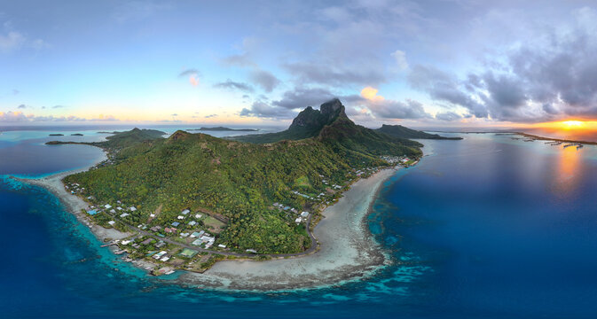 Panoramic aerial view of the tropical island Bora Bora. The sun is coming up over luxury resorts featuring overwater bungalows and huts. The beautiful island is dominated by Mount Otemanu. 