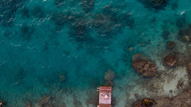 A man laying on his back looking up while on a wooden dock over the south pacific ocean. The sea water is clear and a coral reef is visible underneath the transparent water. 