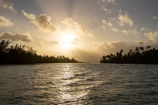 Sunset beyond the water in a channel filled with a coral reef and surrounded by palm trees on the tropical island Taha'a in French Polynesia. 