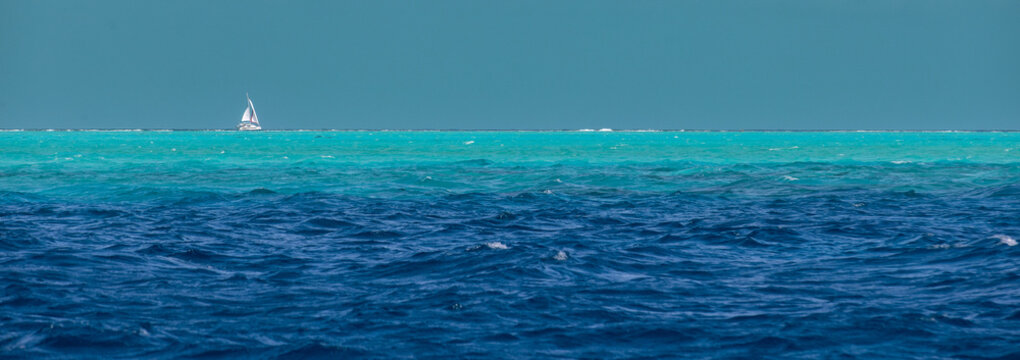 A sailboat can be seen in the distance on the vibrant azure south pacific ocean water sailing over the sea. Beautiful nature shows different shades of blue and turquoise Bora Bora water. .