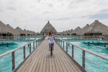 A happy woman rides a bicycle on a wooden dock near overwater bungalows in French Polynesia. The bike ride is over turquoise and azure ocean water on a tropical island motu. 