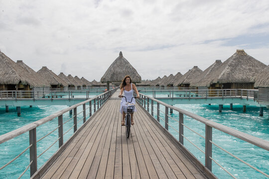 A happy woman rides a bicycle on a wooden dock near overwater bungalows in French Polynesia. The bike ride is over turquoise and azure ocean water on a tropical island motu.  - Powered by Adobe