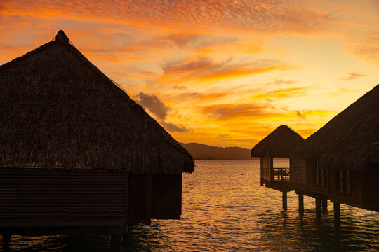 Overwater bungalow huts in silhouette with a vibrant red and orange sunset in the background near tropical island Tahiti or Bora Bora. 