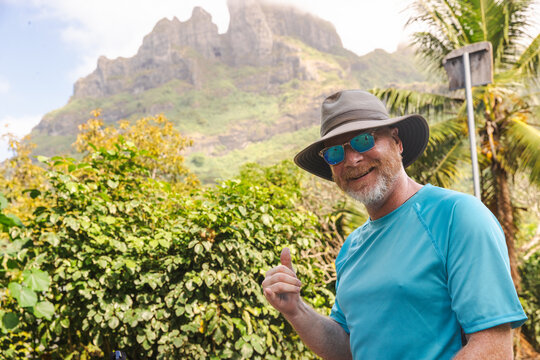 Smiling happy man wearing a floppy hat and sunglasses pointing at Mount Otemanu on Bora Bora island.