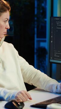 Vertical video Woman listening music while doing software quality assurance, reading source code on computer screen. Developer wearing headphones enjoying podcasts and inspecting coding on desktop PC