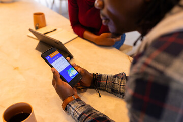 Holding smartphone, African American man checking flight booking app at home with tablet nearby