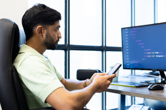 Using smartphone, Indian man sitting at desk with computer screen displaying code