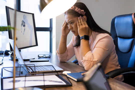Stressed woman in office holding head while working on laptop and documents