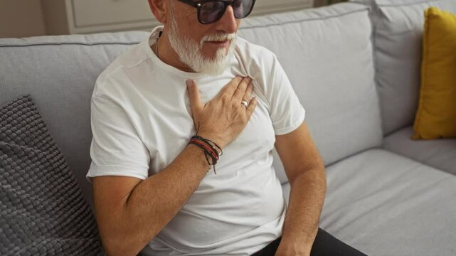 Middle-aged man with a beard and glasses sitting in a living room, touching his chest while seated on a sofa, conveying a sense of concern or discomfort
