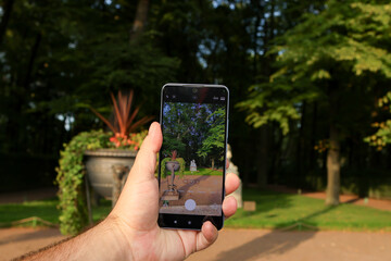 Tourist filming park on cell phone camera. Man's hand holding mobile phone. Guy recording video on smartphone. Male vlogger shoots content in nature. Summer garden and city view