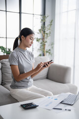 Asian woman holding paper document calculating money savings paying bills in mobile application...