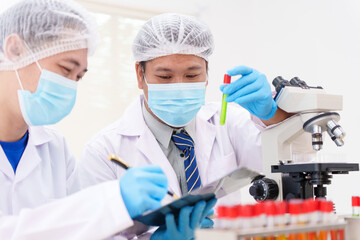 Two Asian scientists in lab coats sit at a table in a laboratory, examining samples through a microscope. The room is equipped with scientific tools, liquids, and chemistry materials.