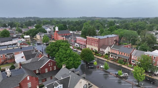 Historic small-town center with brick buildings and tree-lined streets. Main street leads to a church with a white steeple, surrounded by lush greenery. Aerial.