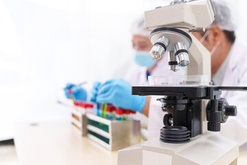 Two Asian scientists in lab coats sit at a table in a laboratory, examining samples through a microscope. The room is equipped with scientific tools, liquids, and chemistry materials.
