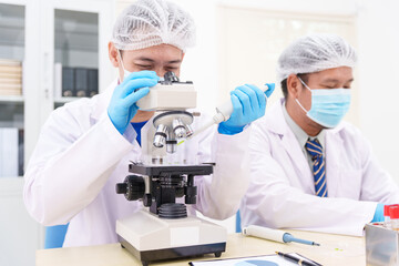 Two Asian scientists in lab coats sit at a table in a laboratory, examining samples through a...