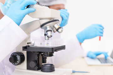 Two Asian scientists in lab coats sit at a table in a laboratory, examining samples through a microscope. The room is equipped with scientific tools, liquids, and chemistry materials.