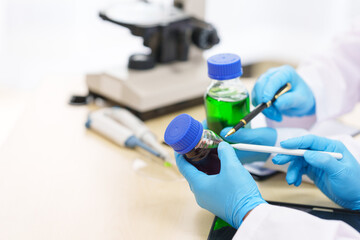 Two Asian scientists in lab coats sit at a table in a laboratory, examining samples through a microscope. The room is equipped with scientific tools, liquids, and chemistry materials.