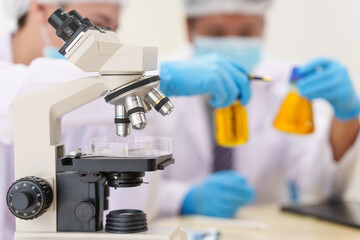 Two Asian scientists in lab coats sit at a table in a laboratory, examining samples through a microscope. The room is equipped with scientific tools, liquids, and chemistry materials.