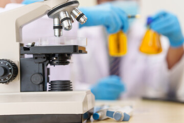 Two Asian scientists in lab coats sit at a table in a laboratory, examining samples through a microscope. The room is equipped with scientific tools, liquids, and chemistry materials.