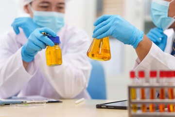 Two Asian scientists in lab coats sit at a table in a laboratory, examining samples through a microscope. The room is equipped with scientific tools, liquids, and chemistry materials.