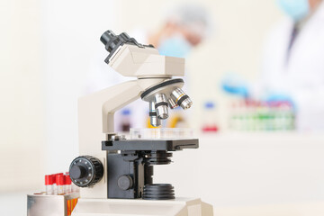 Two Asian scientists in lab coats sit at a table in a laboratory, examining samples through a microscope. The room is equipped with scientific tools, liquids, and chemistry materials.
