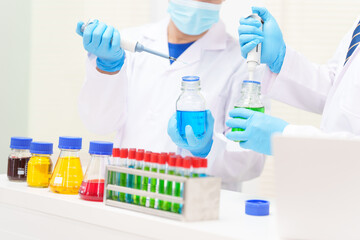 Two Asian scientists in lab coats sit at a table in a laboratory, examining samples through a microscope. The room is equipped with scientific tools, liquids, and chemistry materials.