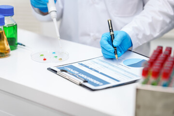 Two Asian scientists in lab coats sit at a table in a laboratory, examining samples through a microscope. The room is equipped with scientific tools, liquids, and chemistry materials.